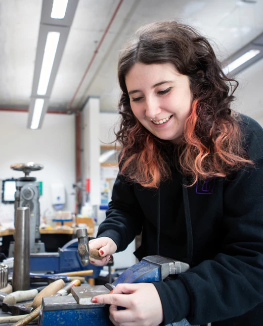A young woman with dark wavy hair, wearing a black hoodie, using a hammer on a jewellery piece at a workbench, surrounded by tools and equipment. A young woman with dark wavy hair, wearing a black hoodie, using a hammer on a jewellery piece at a workbench, surrounded by tools and equipment.